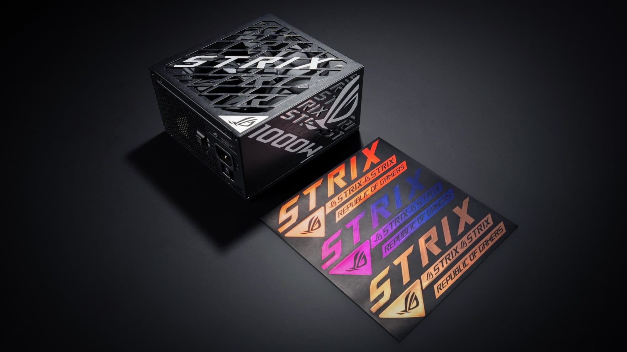 The ROG Strix Platinum 1000W PSU on a gray backdrop beside the gold, purple, and red stickers it comes with