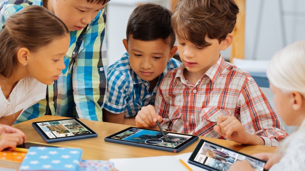 Five young students looking over at one students about to tap on a Lenovo 10w tablet on a table Five young students looking over at one students about to tap on a Lenovo 10w tablet on a table